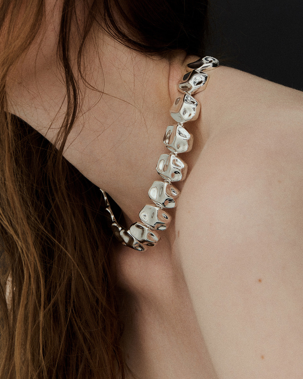 Side profile close-up of a model wearing silver Cosmic Necklace with chunky reflective beads, a bold statement necklace draped along the neck and shoulder, with loose brown hair framing the look.